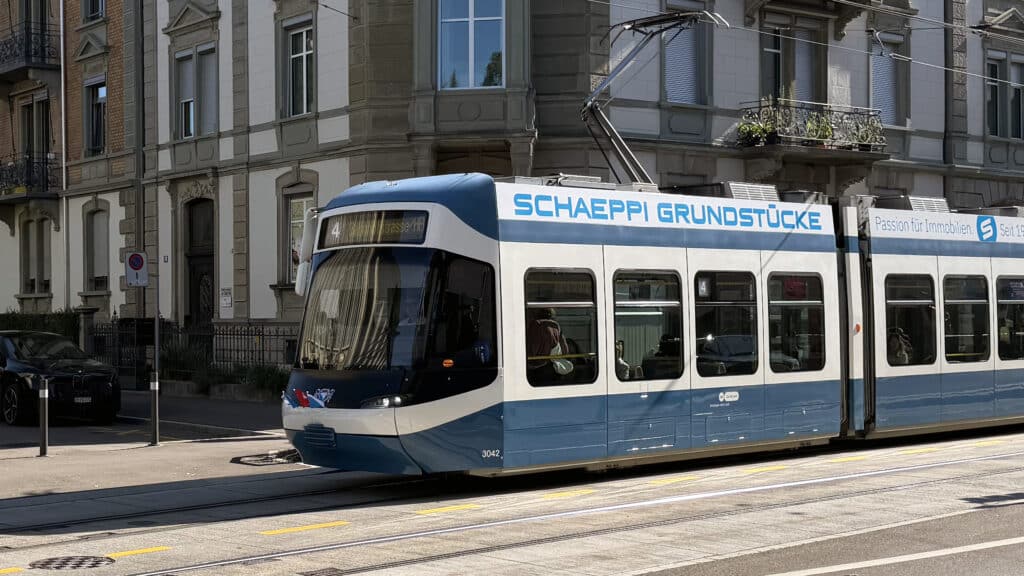 Blau-weisses Cobra-Tram fährt mit kleinen Fahnen an der Front durch eine Zürcher Strasse.