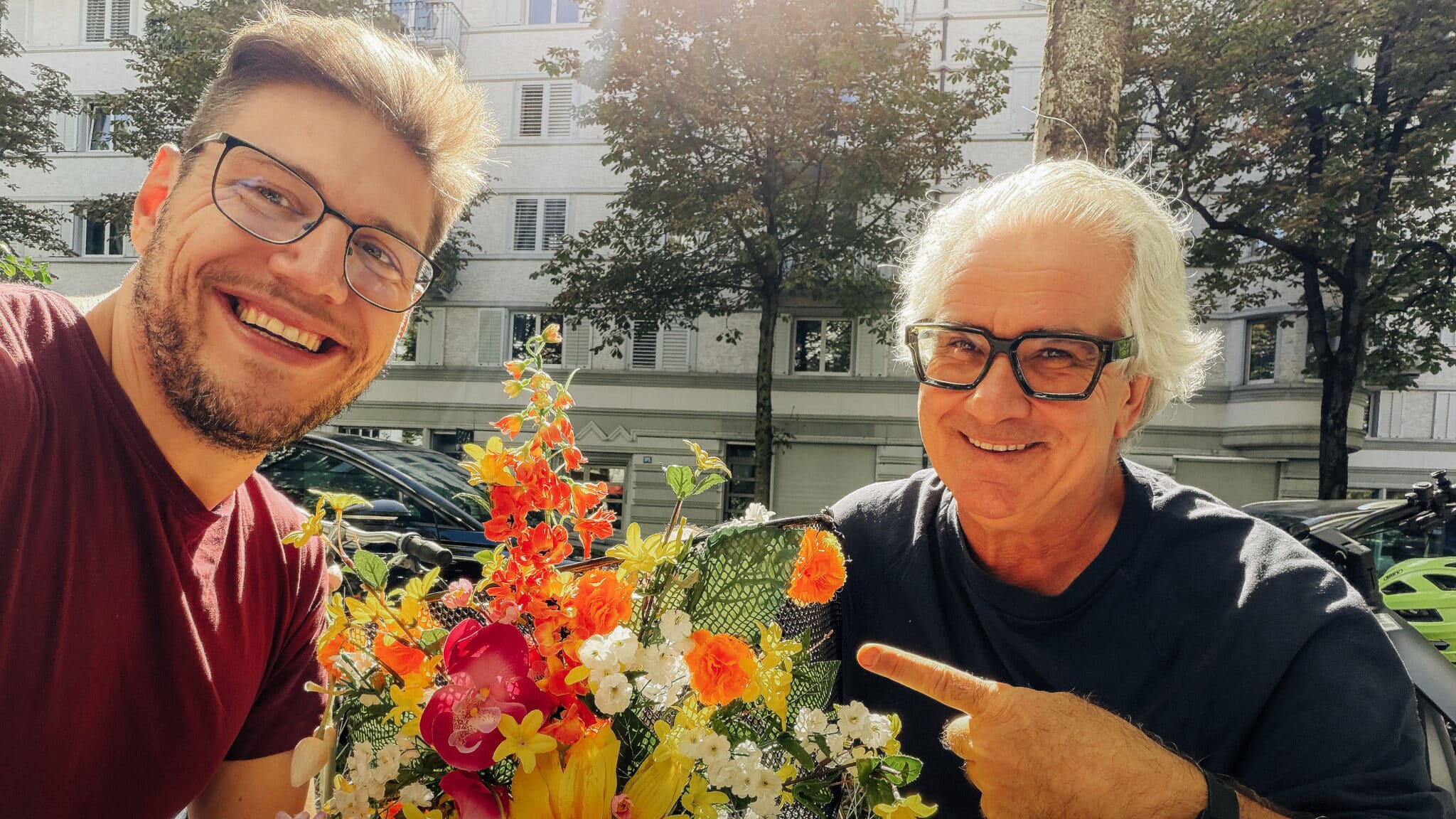 Zwei Männer lächeln in die Kamera, einer zeigt auf einen bunten Blumenstrauss.
