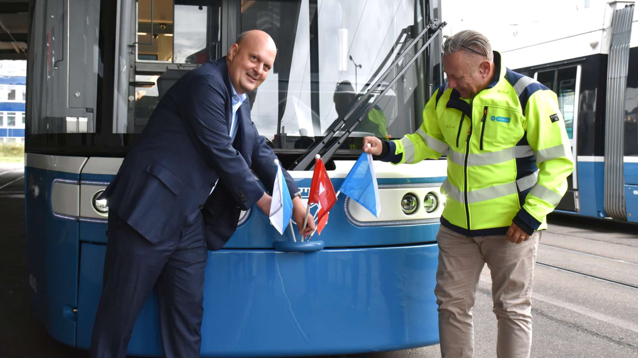 Zwei Männer befestigen kleine Fahnen an der Front eines blau-weissen Flexity-Trams.