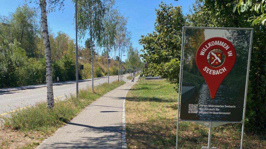 Zu sehen ist eine Strasse mit Trottoir, daneben das Plakat mit dem auffälligen roten Punkt. Die Umgebung ist ländlich und grün.
