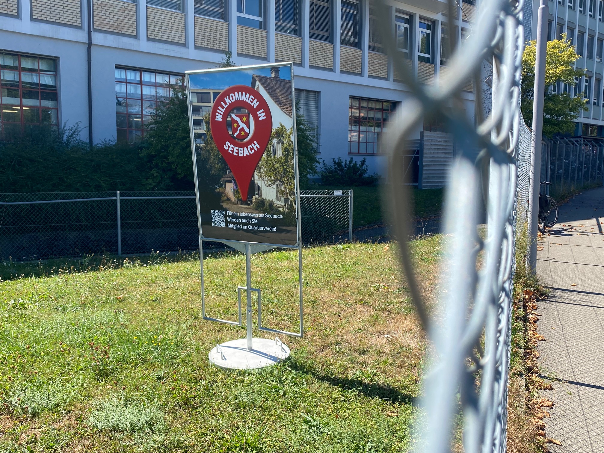 Zu sehen ist ein Plakatständer hinter einem Zaun. Das Suje ist angelehnt an Google Maps mit den charakteristischen roten Punkt. Rechts dominant im Bild der Maschendrahtzaun.