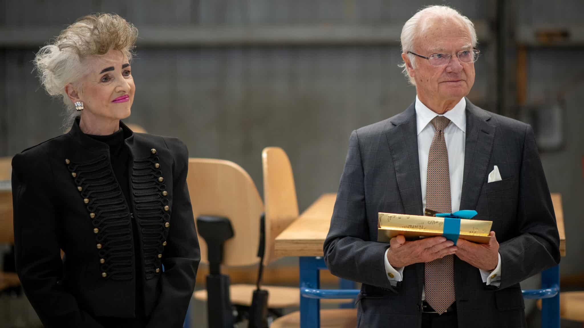 Schwedischer König steht mit einer Zürcher Regierungsrätin in einem Raum und hält ein verpacktes Geschenk mit blauem Band.