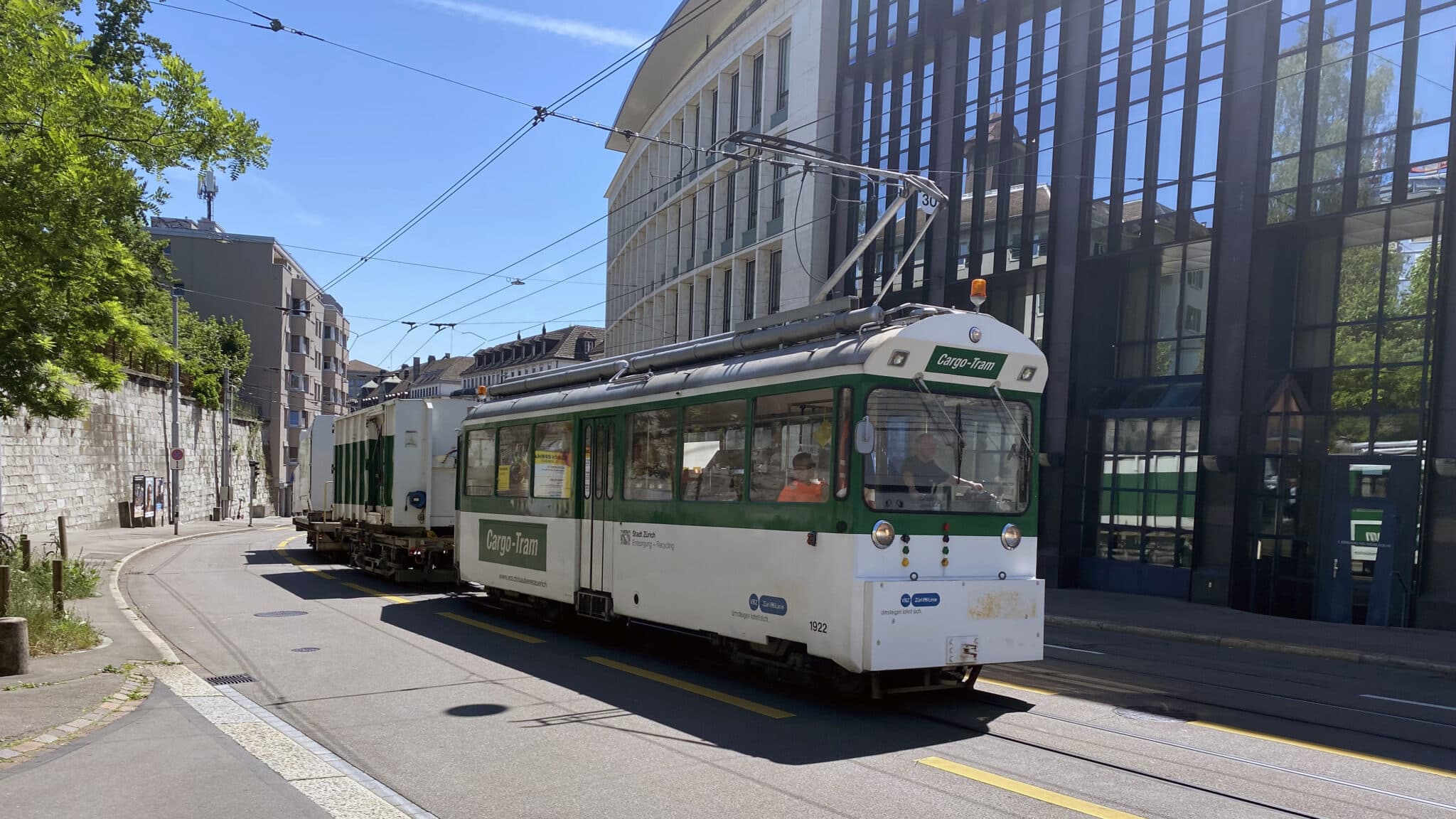 Grün-weisses Cargo-Tram mit angehängtem Recyclingcontainer fährt durch eine sonnige Strasse in Zürich.