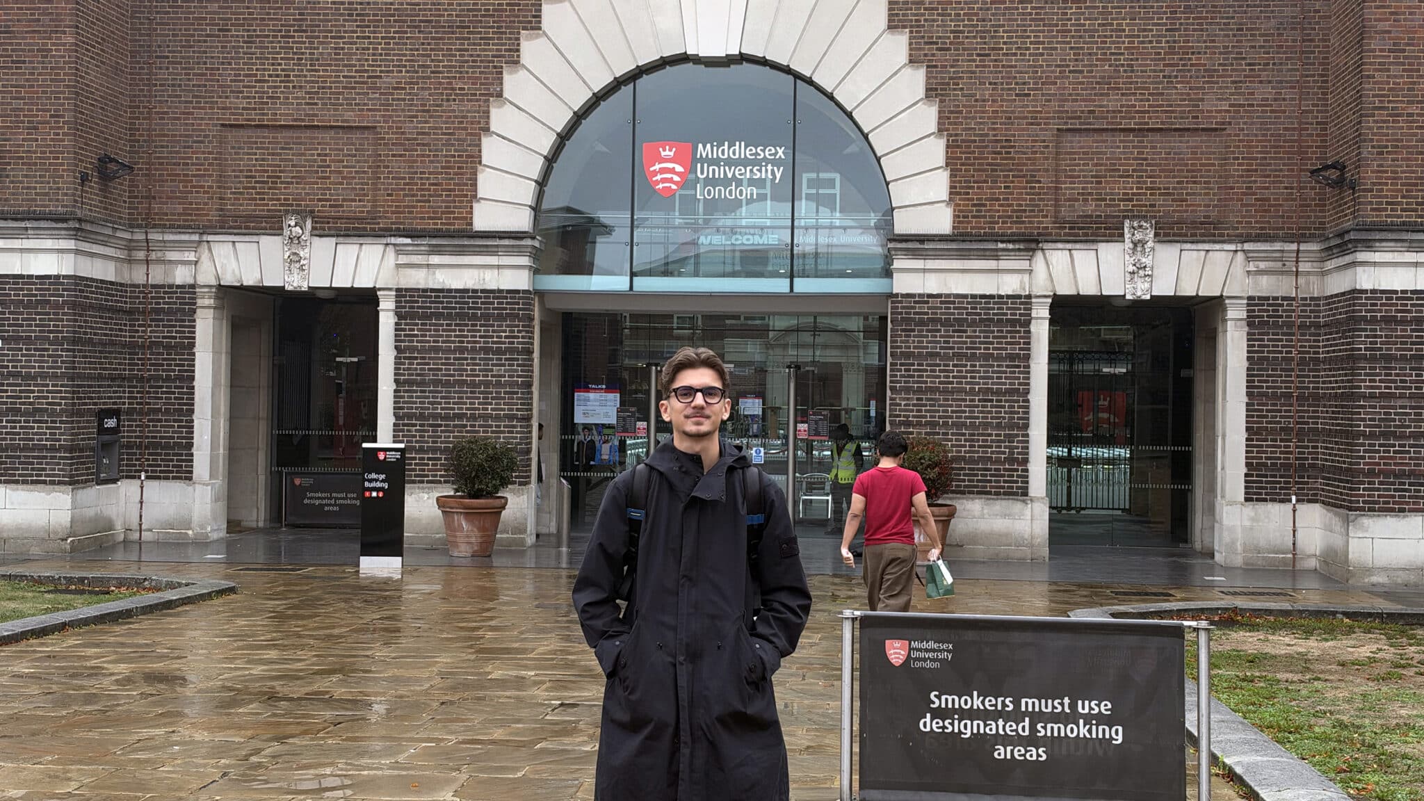 Junger Mann mit Brille und schwarzer Jacke steht vor dem Eingang der Middlesex University in London.