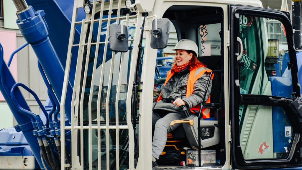 Frau mit weissem Schutzhelm und orangefarbener Sicherheitsweste sitzt lächelnd am Steuer eines Baggers.