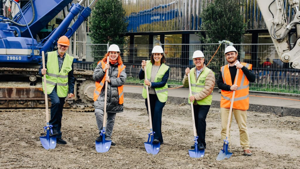 Fünf Personen mit Schutzhelmen und Sicherheitswesten stehen beim symbolischen Spatenstich auf einer Baustelle und halten blaue Schaufeln.