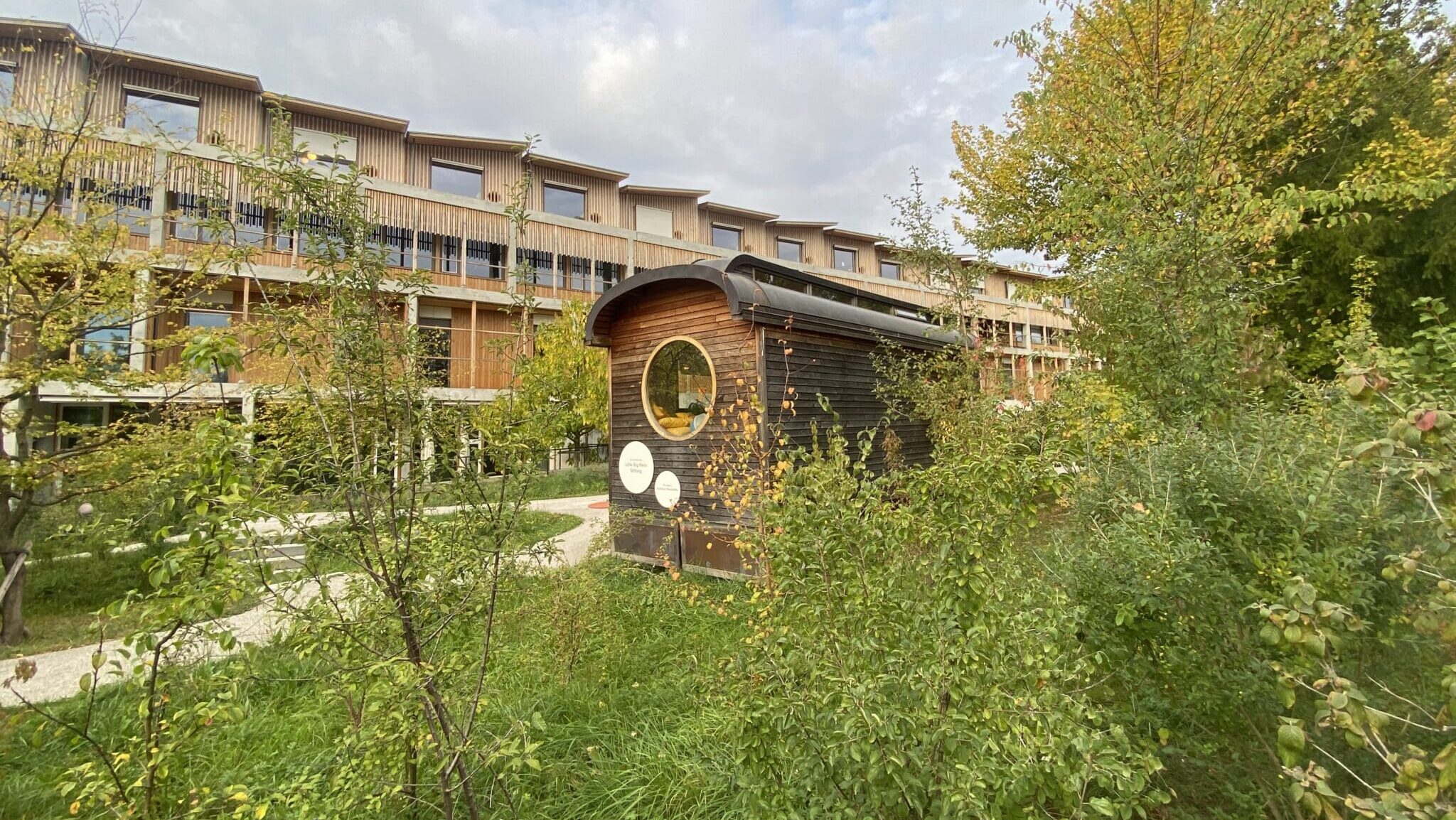 Im Vordergrund steht der kleine Clownwagen aus Holz. Es hat viele Büsche und Bäume rundherum. Im Hintergrund steht das neue Gebäude des Kinderspitals. Die Fassade besteht ebenfalls aus Holz.