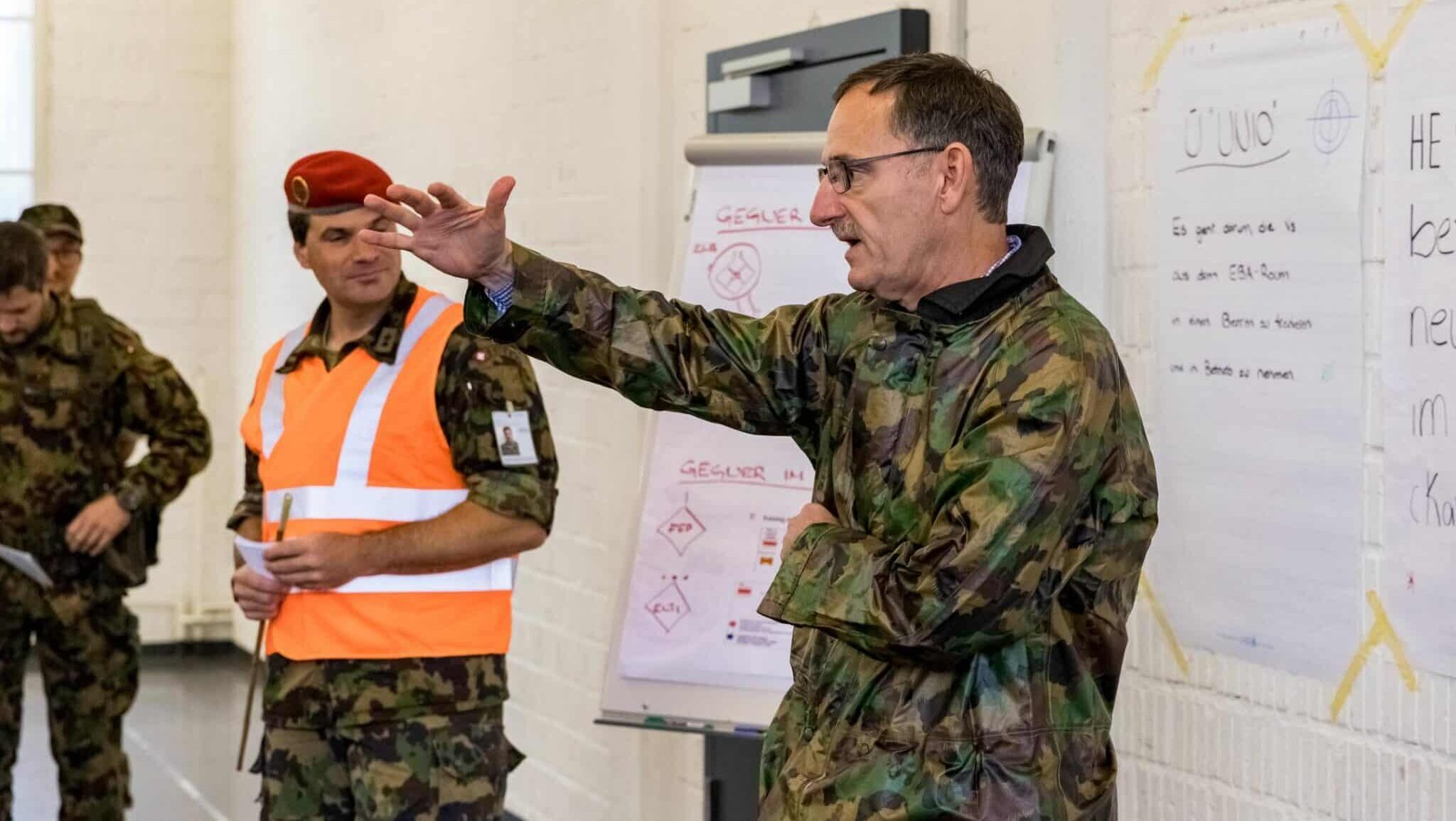 Ein älterer Herr im Tarnanzug hat den rechten Arm leicht ausgestreckt. Ein anderer Man mit einer orangen Weste schaut ihm zu.